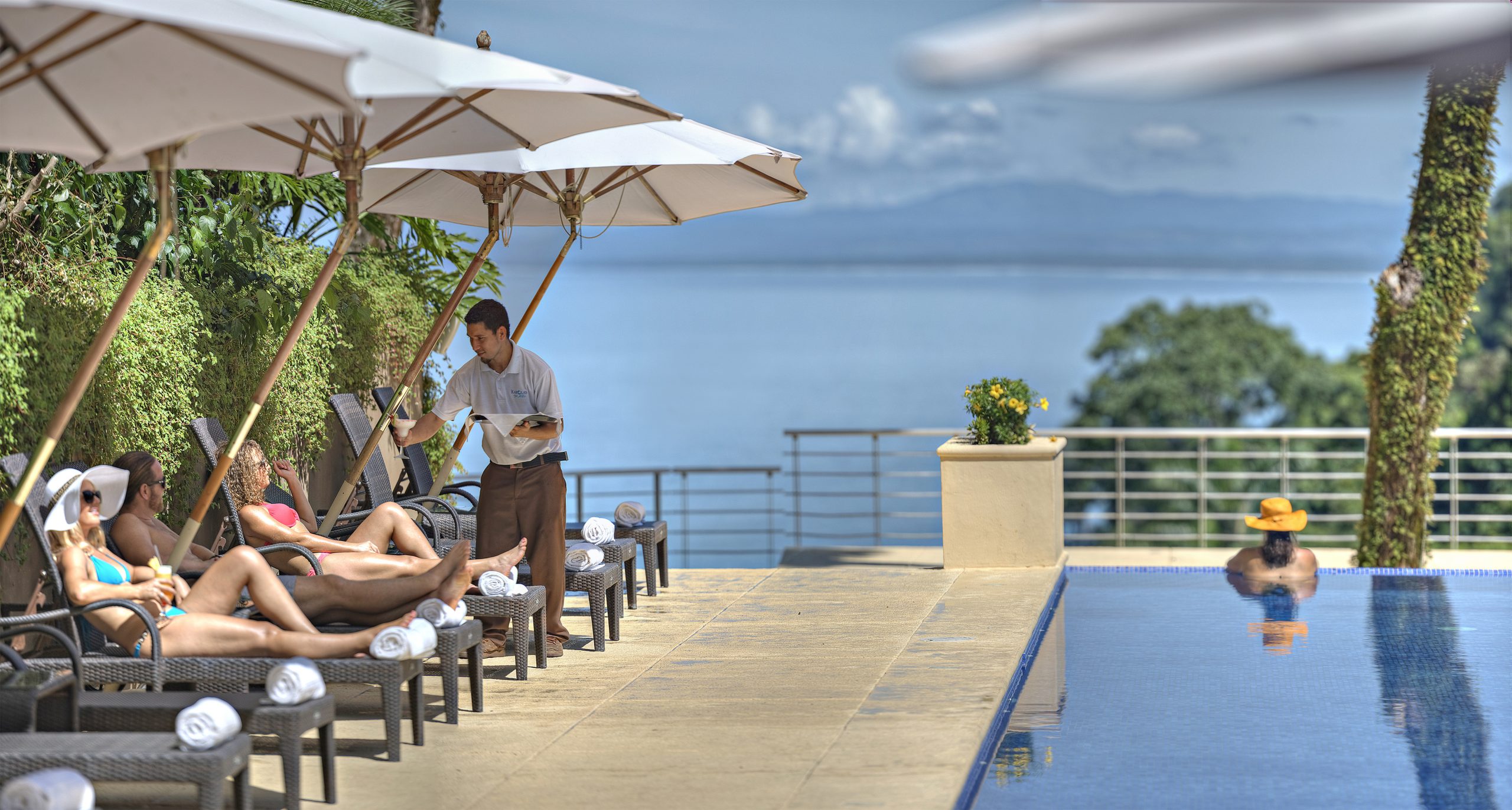 Relaxation by the infinity pool overlooking a serene lake in Costa Rica, with guests sunbathing and a server attending to their needs.