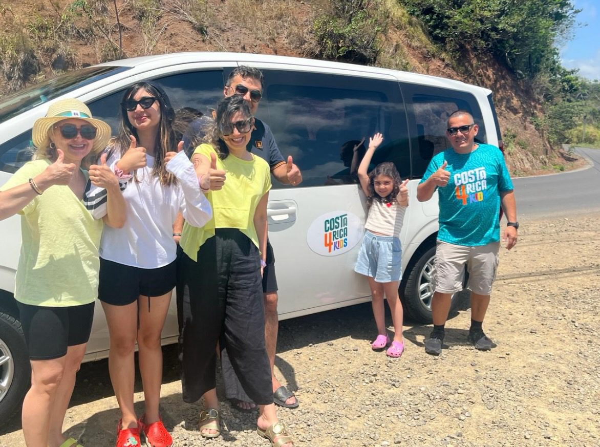 A joyful group of tourists giving thumbs up next to a van with "Costa Rica For Kids" written on it, indicating they're having a great time on their trip.