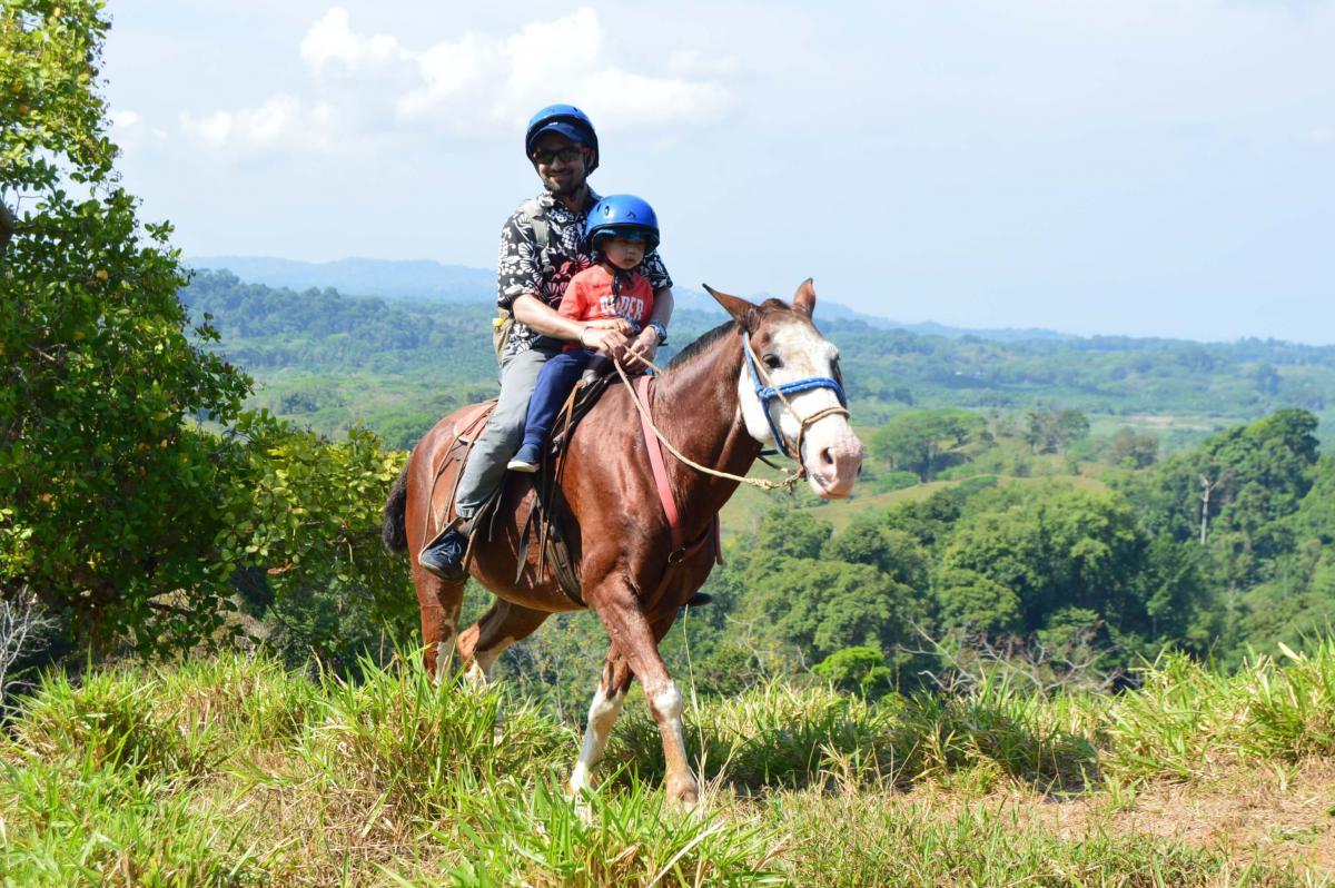 A joyful horseback riding adventure in Costa Rica, featuring an adult and a child, both wearing helmets, on a scenic trail with lush greenery and a clear blue sky in the background.