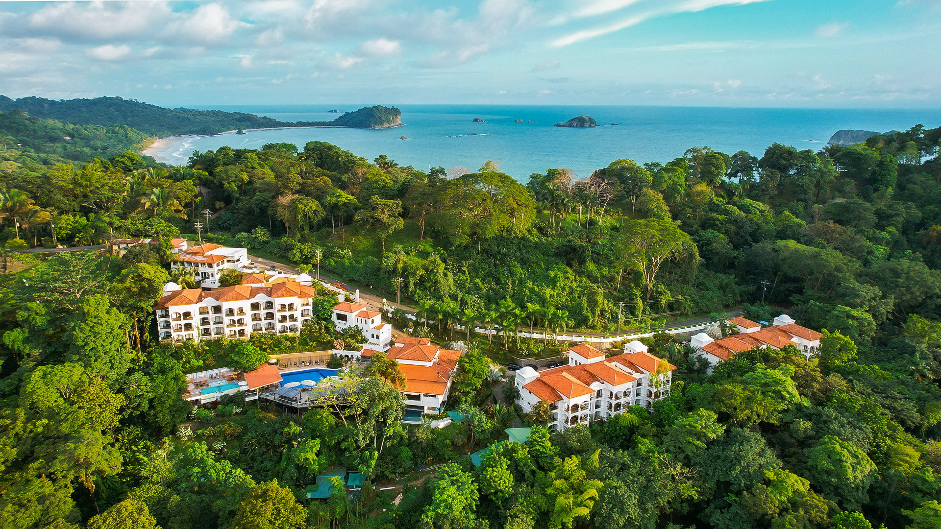 Aerial view of a tropical resort nestled in lush greenery with a view of the ocean and distant islands.