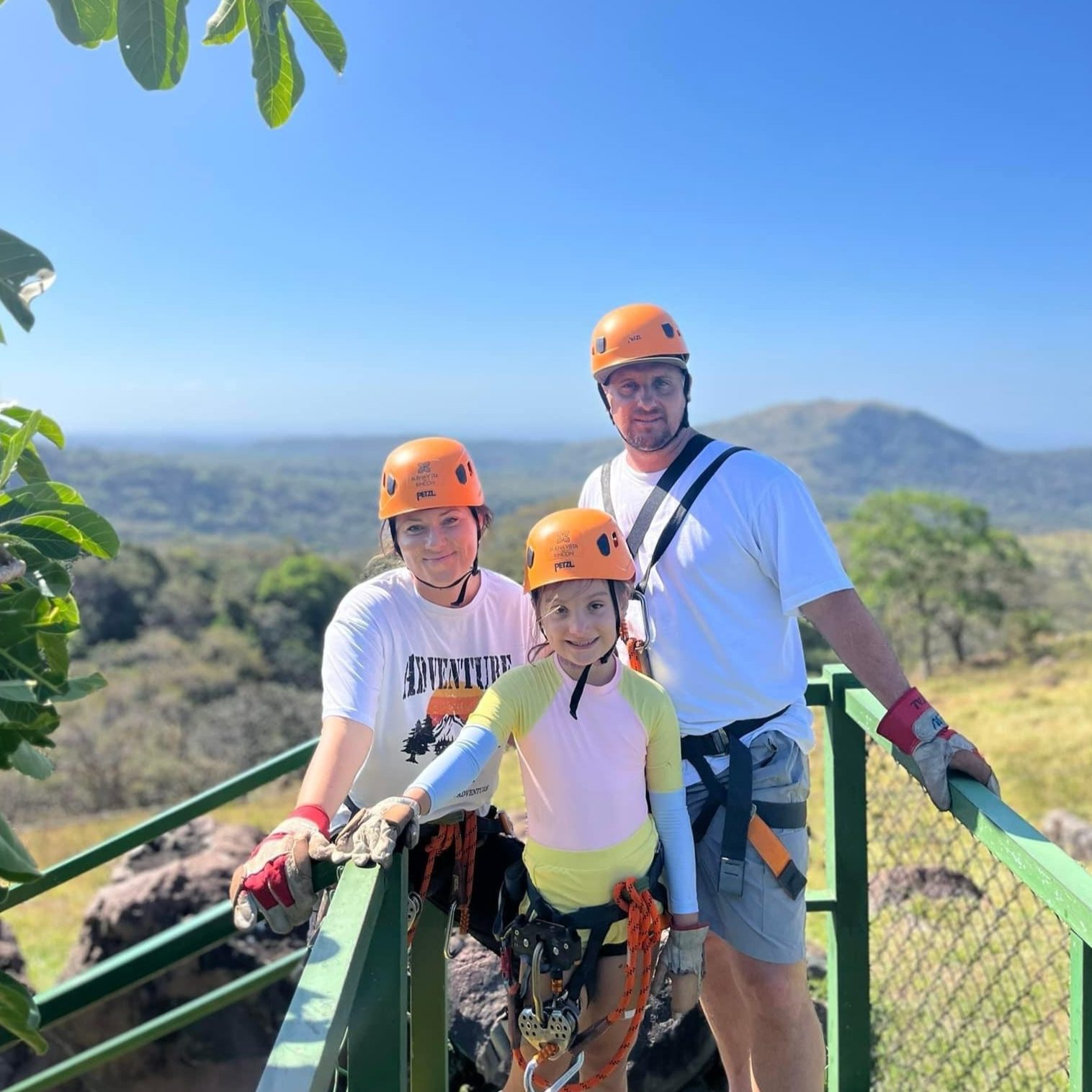 A family dressed in outdoor adventure gear with helmets and harnesses, ready for a thrilling zipline experience in Costa Rica, boasting lush greenery and rolling hills perfect for kids.
